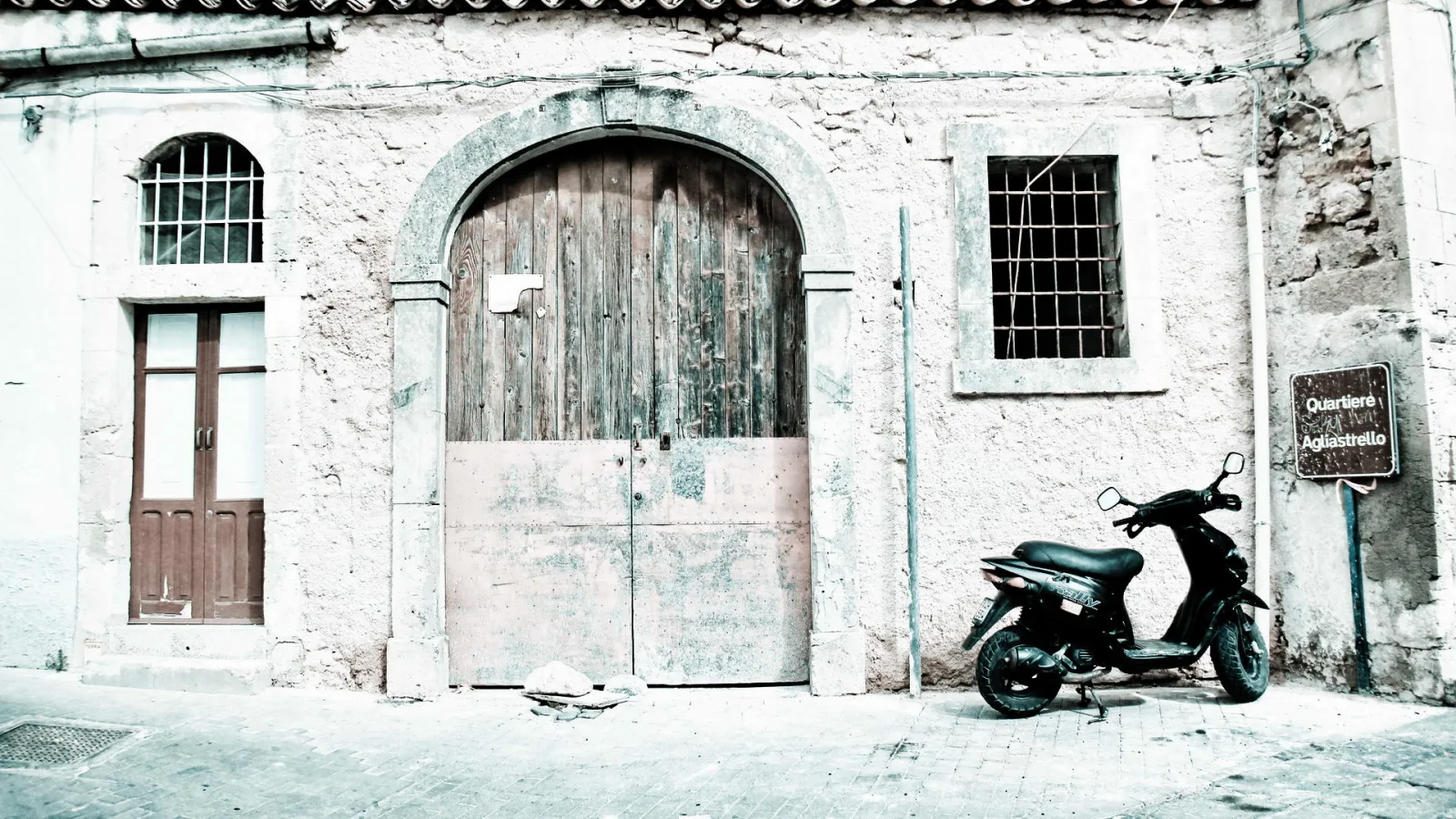 Verwitterte Steinfassade mit großem Rundbogentor aus dunklem Holz, links eine Holztür, rechts ein vergittertes Fenster, davor ein abgestellter schwarzer Motorroller auf gepflastertem Platz.