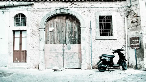 Verwitterte Steinfassade mit großem Rundbogentor aus dunklem Holz, links eine Holztür, rechts ein vergittertes Fenster, davor ein abgestellter schwarzer Motorroller auf gepflastertem Platz.