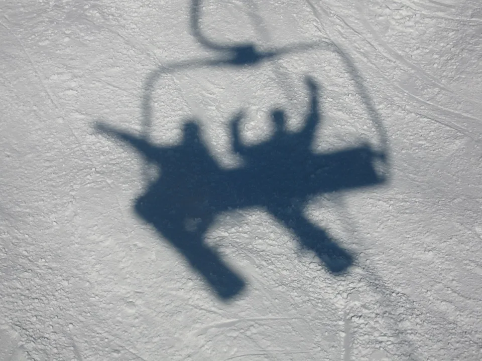 Schatten eines Sessellifts mit zwei Personen auf einer hellen Schneefläche