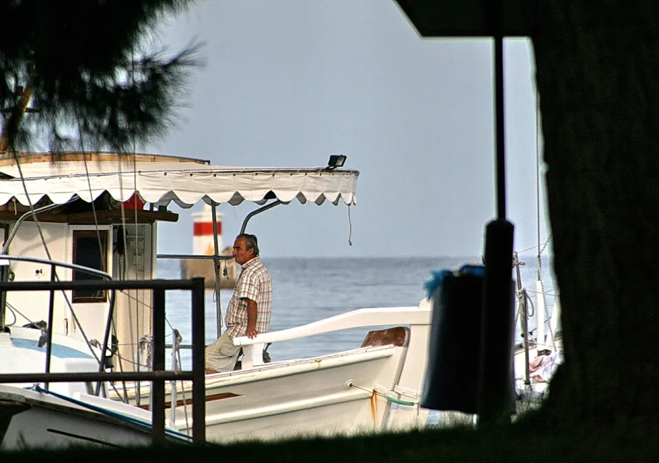 Ein Mann mit kariertem Hemd steht auf dem Deck eines weißen Bootes, dahinter Meer und ein Leuchtturm mit roten Streifen.