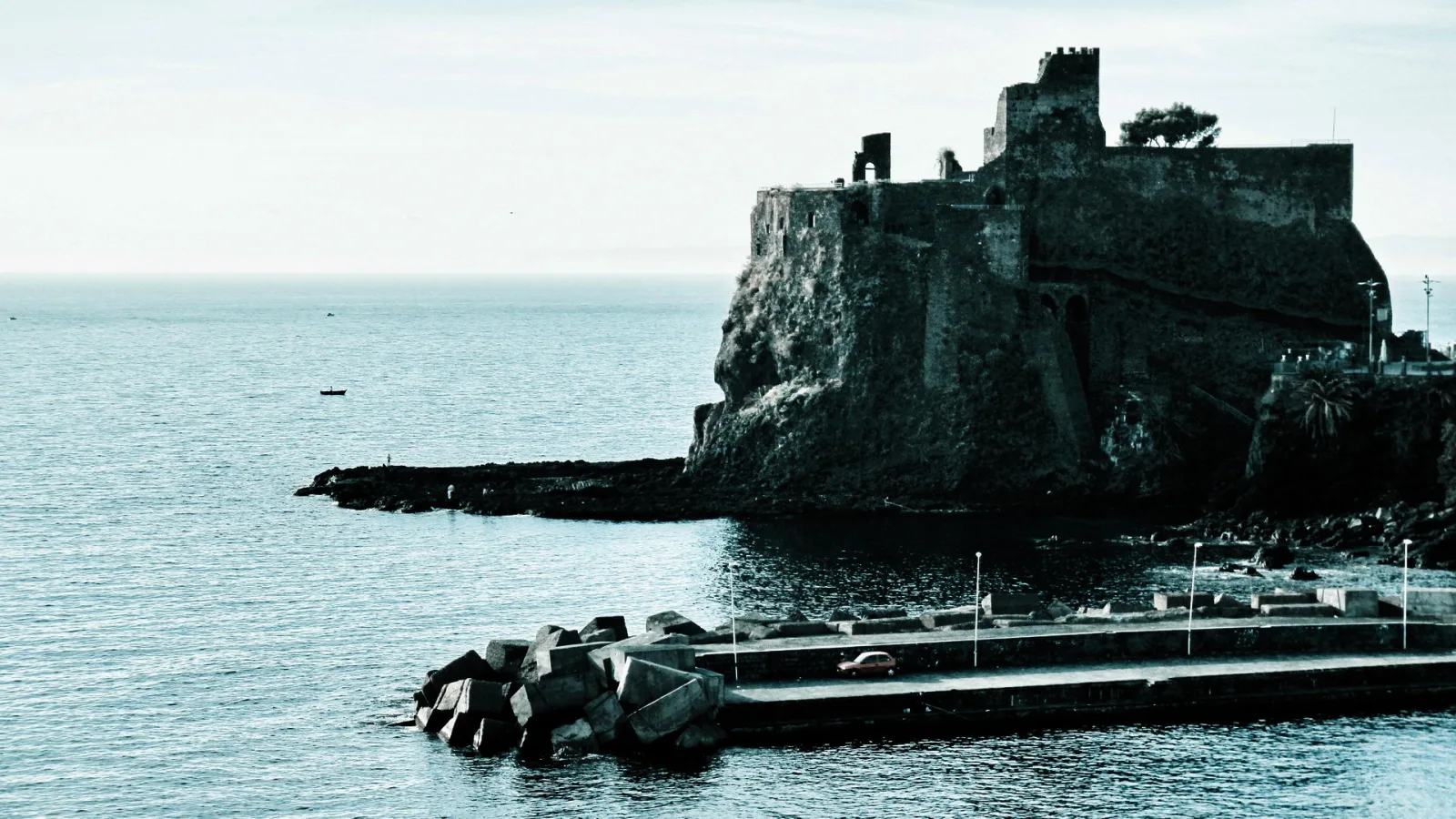 Mittelalterliche Burg auf einem Felsen am Meer, davor eine Mole mit Betonblöcken und ein kleines Boot auf dem Wasser