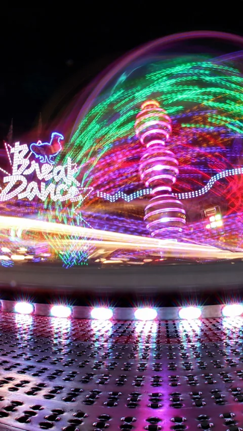 Fahrgeschäft auf einem Jahrmarkt bei Nacht, bunte Lichtspuren und eine leuchtende Spirale, im Vordergrund eine glänzende Plattform mit Lampen, links der Schriftzug Break Dance.