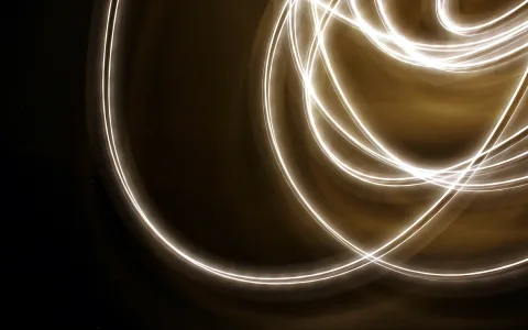 Mehrere helle, geschwungene Lichtspuren auf dunklem Grund, die sich überkreuzen und nach rechts oben zu einem dichten Knäuel werden, von warmem Beige-Braun umgeben.
