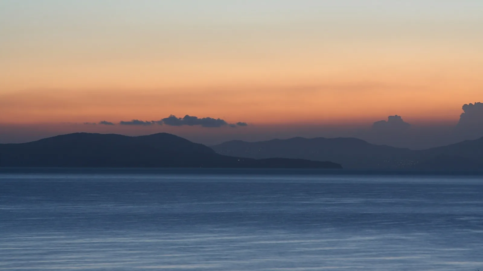 Dunkle Inseln und Bergrücken am Horizont über glattem blauem Meer, darüber ein orange-blauer Abendhimmel mit wenigen Wolken.