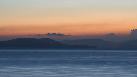 Dunkle Inseln und Bergrücken am Horizont über glattem blauem Meer, darüber ein orange-blauer Abendhimmel mit wenigen Wolken.
