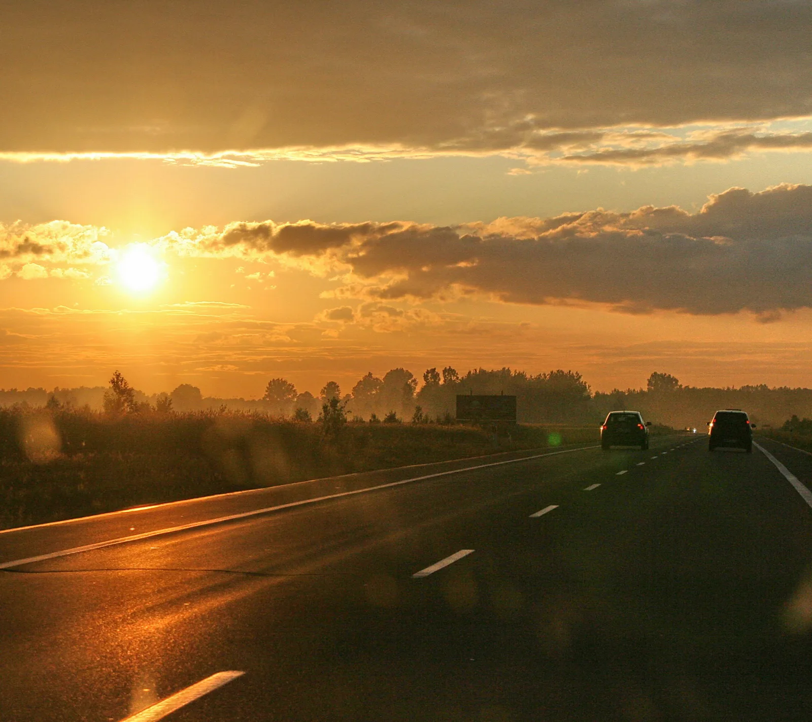 Asphaltierte Autobahn mit weißen Fahrbahnmarkierungen führt auf eine tief stehende Sonne zu, zwei Autos fahren in dieselbe Richtung, am Horizont Baumreihen und Dunst, der Himmel ist orange und von Wolken durchzogen.