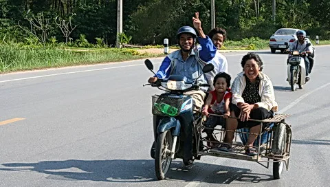 Ein Mann fährt einen Motorroller mit Beiwagen auf einer sonnigen Straße. Im Beiwagen sitzen ein Kind und eine lächelnde Frau, hinter dem Fahrer winkt ein Mann, und im Hintergrund fährt ein Motorroller vorbei.