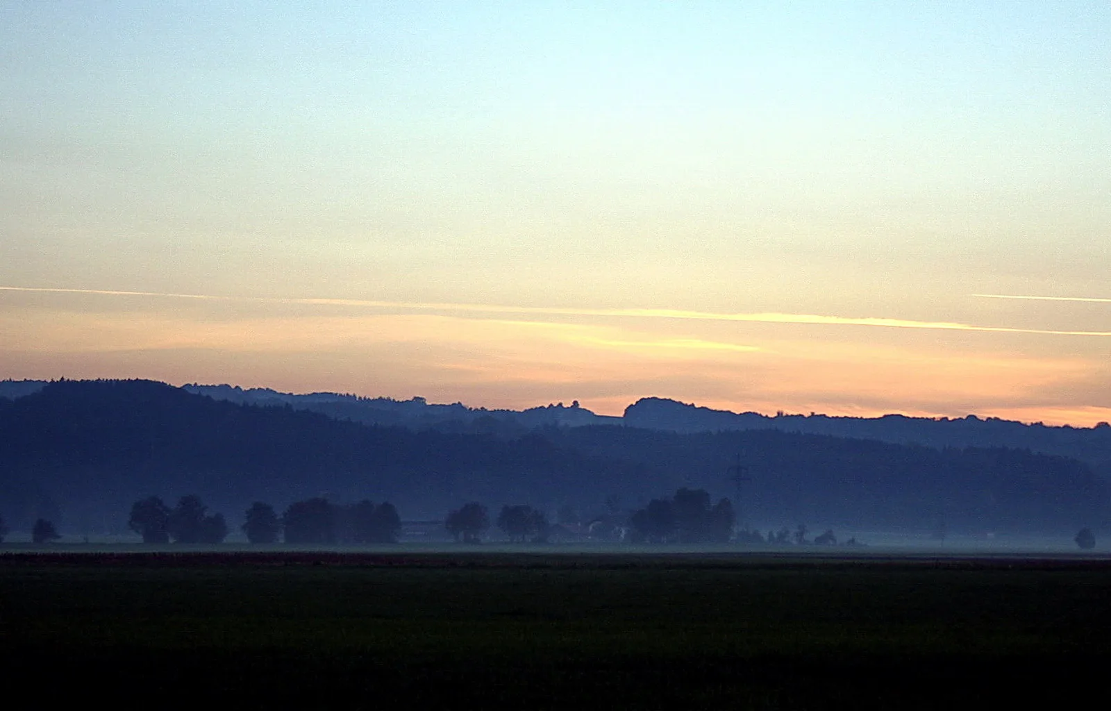 Weite Landschaft im Dämmerlicht: dunkle Hügelkämme und Baumgruppen im Dunst, darüber ein Himmel in Blau und Orange mit feinen Kondensstreifen, im Vordergrund eine dunkle Ackerfläche.