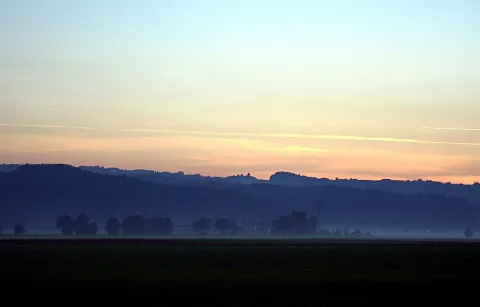 Weite Landschaft im Dämmerlicht: dunkle Hügelkämme und Baumgruppen im Dunst, darüber ein Himmel in Blau und Orange mit feinen Kondensstreifen, im Vordergrund eine dunkle Ackerfläche.