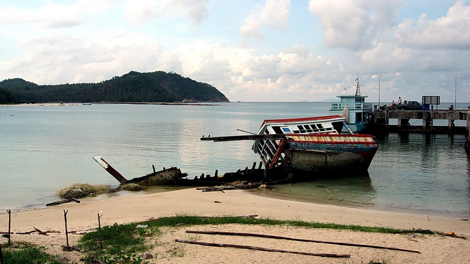 Ein halb gesunkenes, verrostetes Boot mit bunt gestrichenem Rumpf liegt auf einem Sandstrand. Dahinter liegen ein Anleger mit Gebäude und ein Boot im ruhigen Wasser, links bewaldete Hügel unter bewölktem Himmel.