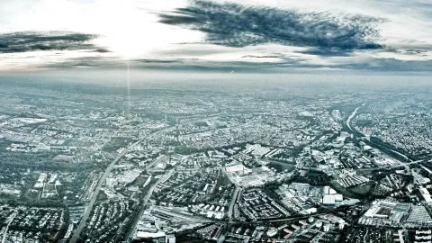 Luftaufnahme einer Stadt mit Straßen, Häuserdächern und Industriegebäuden unter einem bewölkten Himmel, am Horizont liegt Dunst.