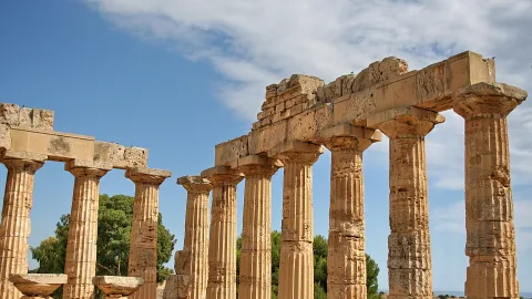 Mehrere beige Säulen mit Kapitellen eines antiken griechischen Tempels vor blauem Himmel mit Wolken, im Hintergrund grüne Bäume.