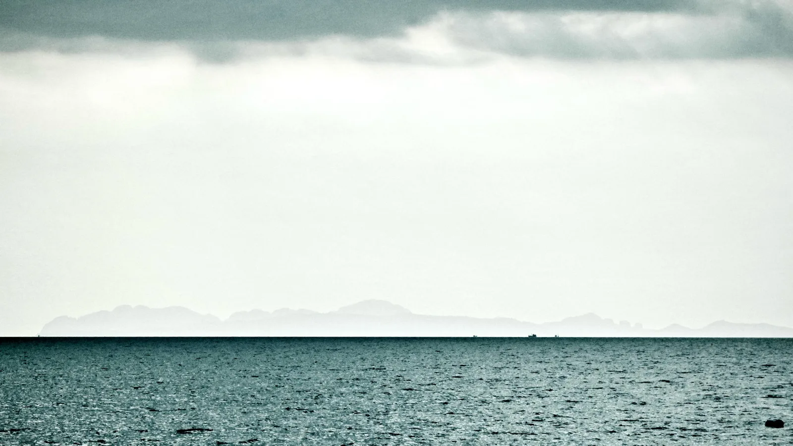 Ruhiges, dunkelblaues Meer unter einem bewölkten, hellgrauen Himmel. Am Horizont zeichnen sich blasse Inselkonturen ab, auf dem Wasser sind zwei winzige Schiffe zu erkennen.