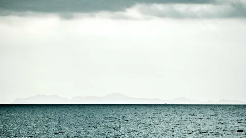 Ruhiges, dunkelblaues Meer unter einem bewölkten, hellgrauen Himmel. Am Horizont zeichnen sich blasse Inselkonturen ab, auf dem Wasser sind zwei winzige Schiffe zu erkennen.