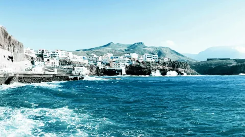 Weiß getünchte Häuser auf einer Klippe über dem blauen Meer, davor brechen Wellen an einer Hafenmauer, dahinter ragt ein Berg auf.