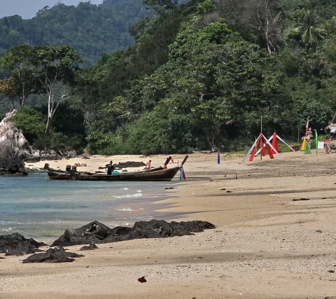 Ein Langschwanzboot liegt am Sandstrand vor dunklen Felsen, dahinter steigt dichter grüner Wald an, und rechts stehen bunte Fahnen.