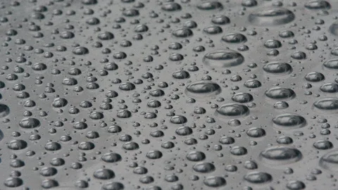 Viele kleine und größere Wassertropfen liegen dicht verteilt auf einer grauen, glatten Oberfläche.