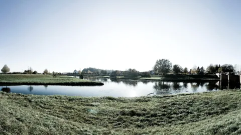 Breiter Fluss mit grasbewachsenem Damm, Brücke und Schwänen auf dem Wasser unter hellem Himmel.