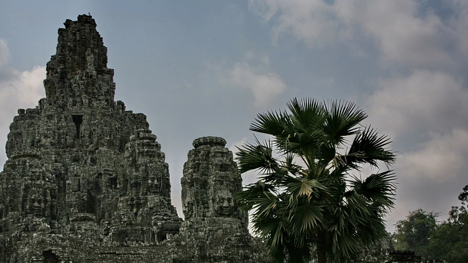 Verwitterte Steintürme des Bayon-Tempels mit großen Gesichtsreliefs, rechts davon eine Fächerpalme vor bewölktem Himmel.