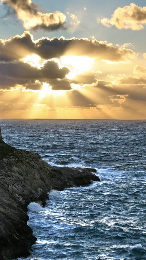 Felsige Küste im Vordergrund, dahinter aufgewühltes Meer und tiefer Horizont, darüber Wolken, durch die die tief stehende Sonne goldene Strahlen wirft.