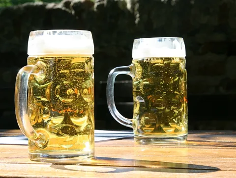 Zwei Biergläser mit hellem Bier und Schaumkrone stehen auf einem Holztisch vor einer dunklen Steinmauer.
