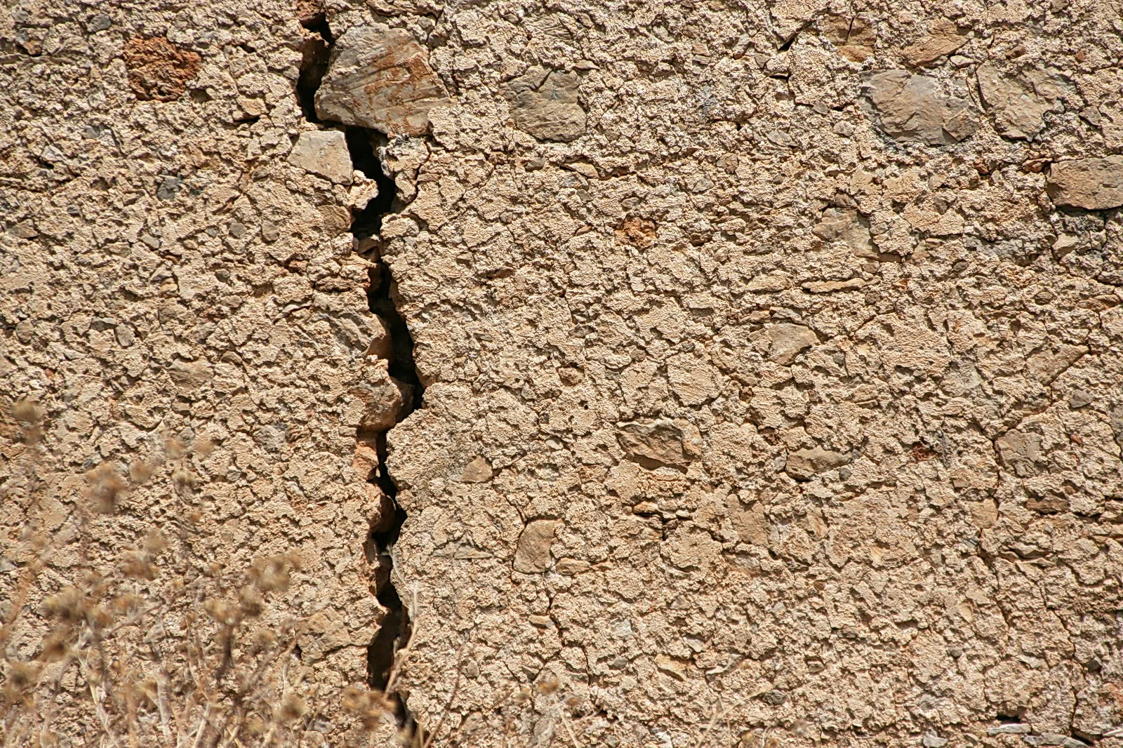 Beigebraune, rissige Mauer aus Putz und eingelassenen Steinen, ein tiefer vertikaler Riss verläuft durch das Bild, unten links trockene Grashalme.
