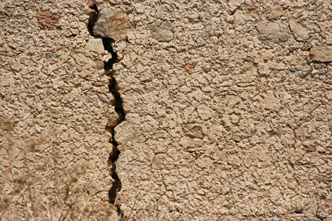Beigebraune, rissige Mauer aus Putz und eingelassenen Steinen, ein tiefer vertikaler Riss verläuft durch das Bild, unten links trockene Grashalme.