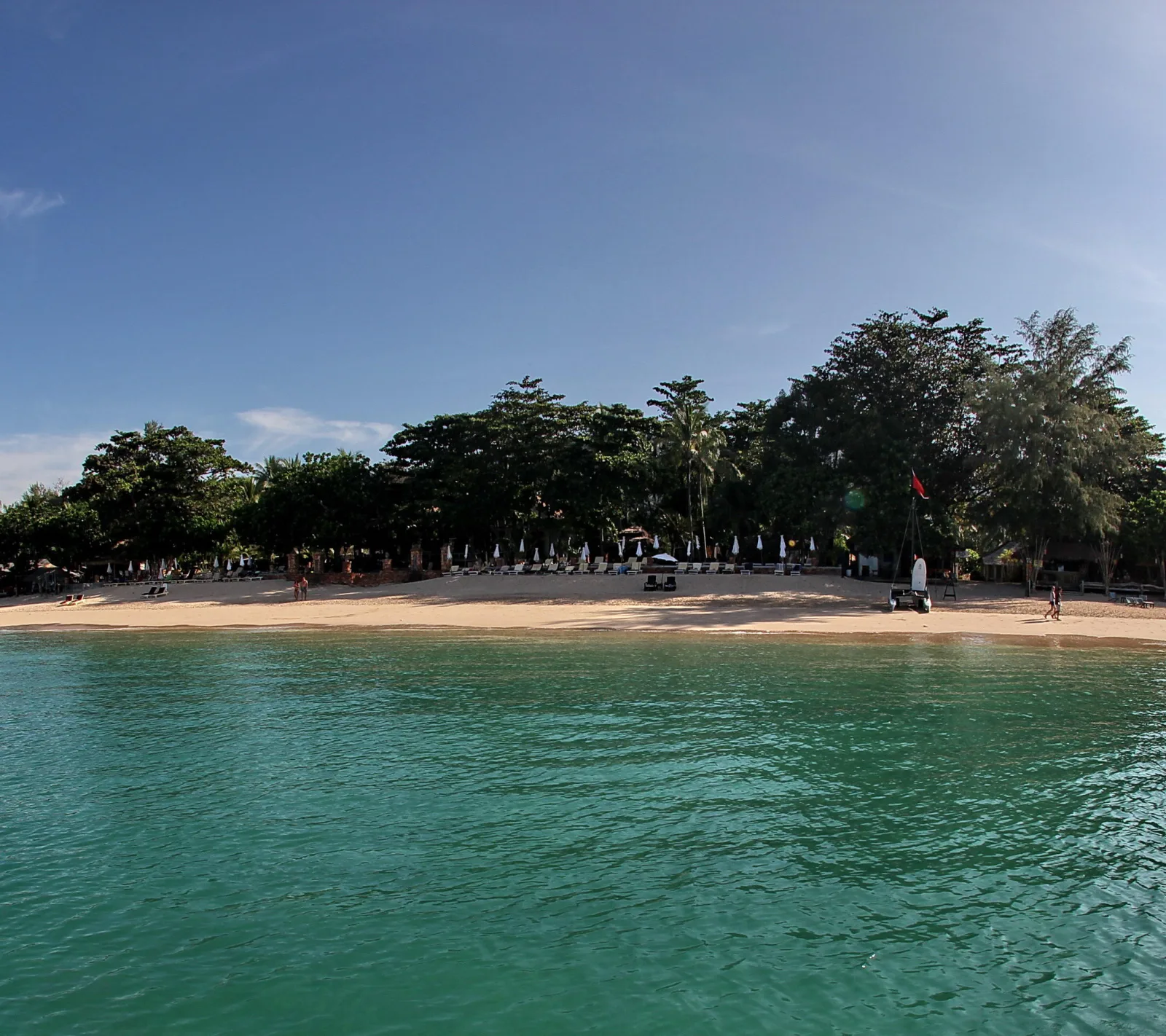 Sandstrand mit Sonnenliegen und Sonnenschirmen vor dichten grünen Bäumen, davor klares türkisfarbenes Meer unter blauem Himmel
