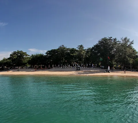 Sandstrand mit Sonnenliegen und Sonnenschirmen vor dichten grünen Bäumen, davor klares türkisfarbenes Meer unter blauem Himmel