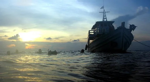 Ein Holzboot liegt am Abend auf dem Meer, links schwimmen mehrere Taucher im Wasser, im Hintergrund geht die Sonne hinter Wolken unter.