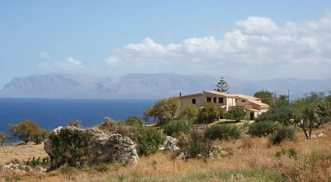 Ein hellgelbes Landhaus mit Ziegeldach auf einem trockenen Hügel, links das blaue Meer, dahinter ein langer Bergrücken unter Wolken.