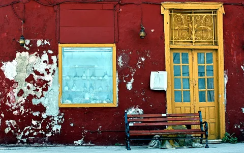 Rot verputzte Hauswand mit abblätternem Putz, links ein Fenster mit gelbem Rahmen und Flaschen dahinter, rechts eine gelbe Holztür mit verschnörkeltem Gitter, davor eine Holzbank und ein liegender Sack auf dem Gehweg.