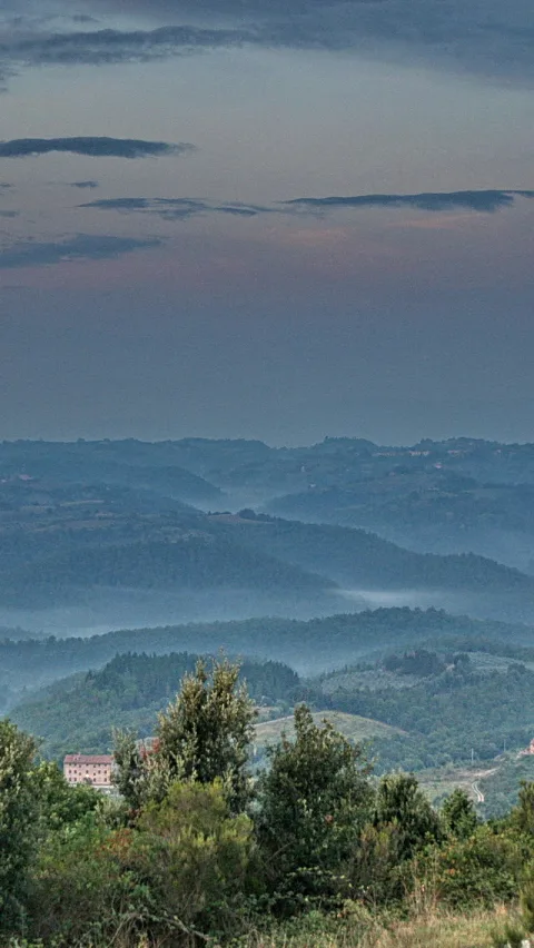 Weite Hügellandschaft in Umbrien mit blauem Morgennebel in den Senken, im Vordergrund grüne Bäume und ein rosa Gebäude am Hang, darüber ein blassblauer Himmel mit länglichen Wolken.