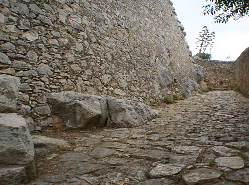 Schmale Gasse mit grobem Kopfsteinpflaster, links eine hohe Mauer aus Bruchsteinen, im Hintergrund eine Agave und heller Himmel.