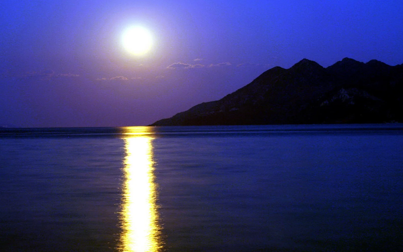 Heller Vollmond über einem ruhigen, blauen Meer am Abend, dessen Spiegelung als gelbe Bahn zum Ufer führt; rechts ein dunkler Gebirgszug.