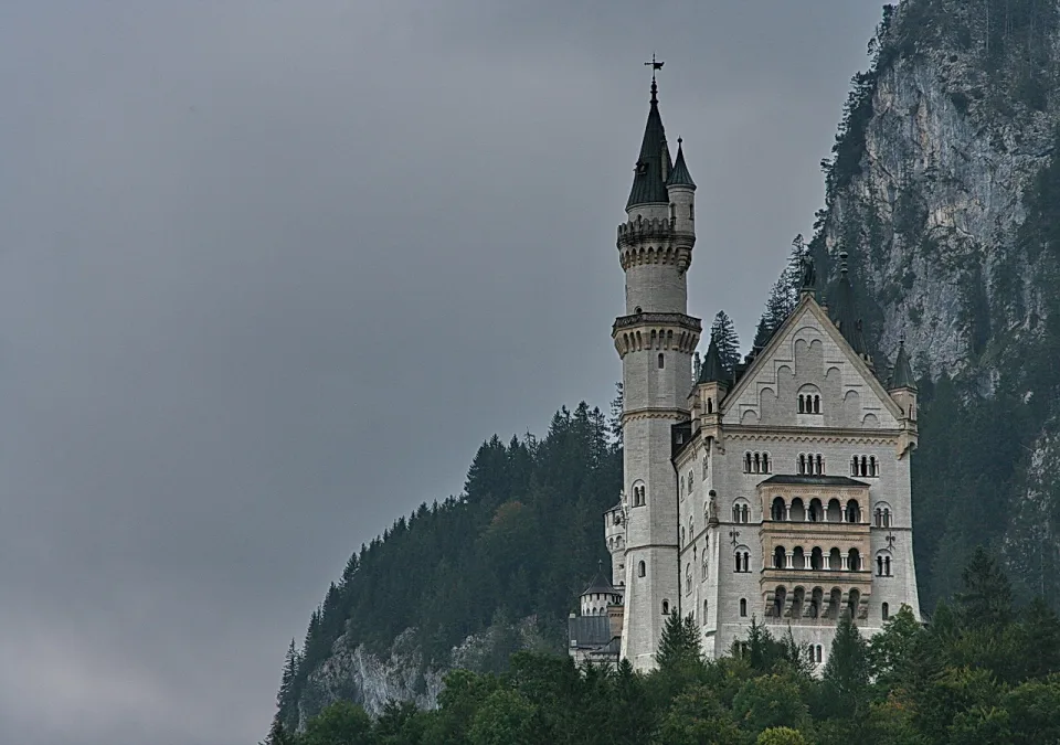 Das weiße Schloss Neuschwanstein mit spitzen Türmen auf einem bewaldeten Felsen unter grauem Himmel.