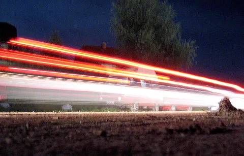 Langzeitbelichtung einer nächtlichen Straße mit roten und weißen Lichtspuren vorbeifahrender Autos, dahinter eine Baumgruppe und dunkler Himmel.