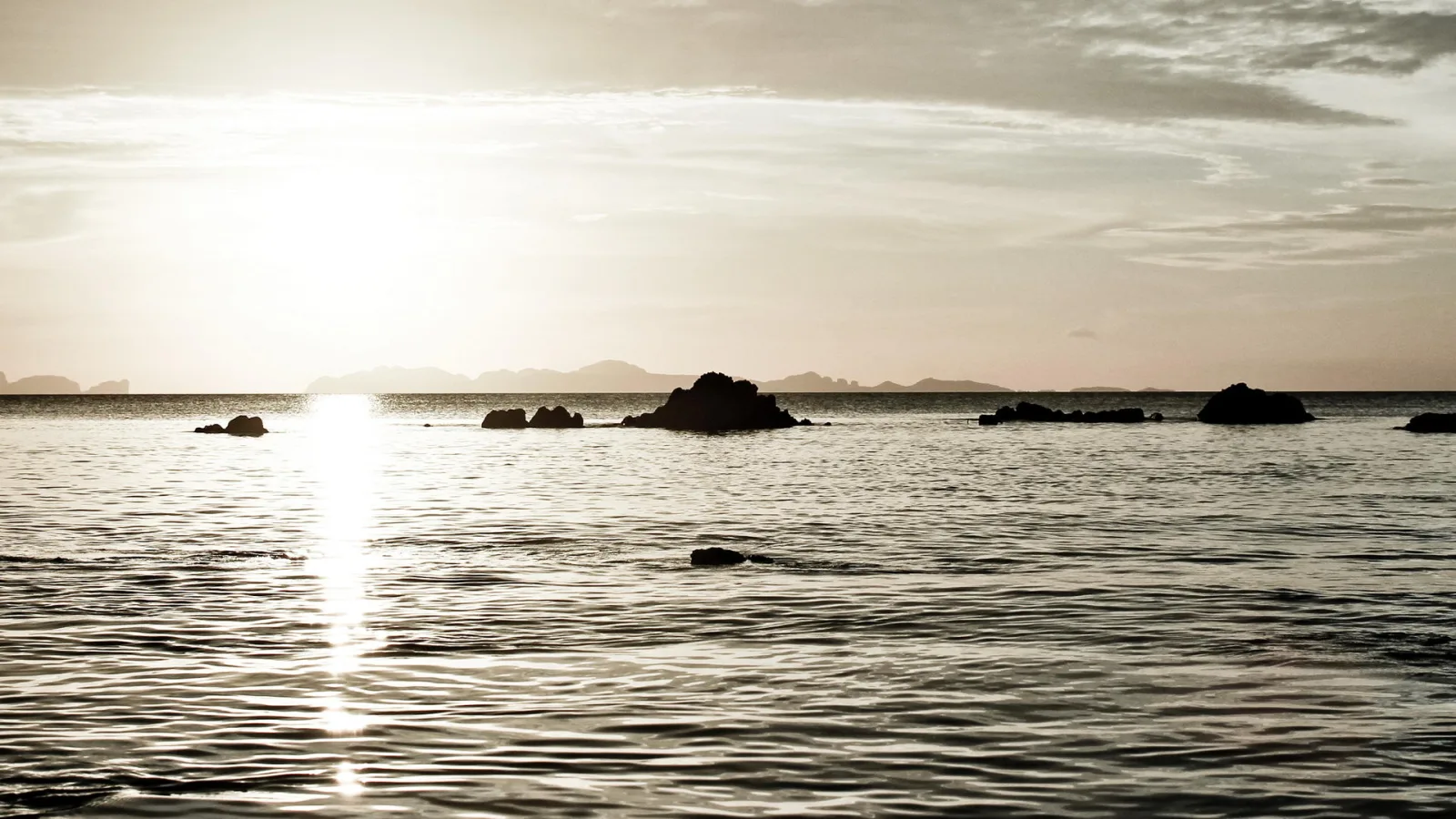 Ruhiges Meer im warmen Gegenlicht der tiefstehenden Sonne, aus dem Wasser ragen mehrere dunkle Felsen und Inseln, im Hintergrund zeichnen sich flache Bergketten am Horizont ab.