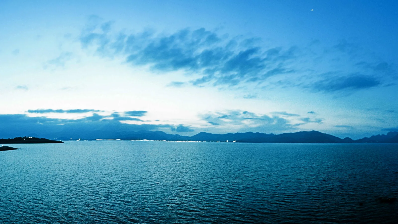 Ruhiges Meer unter blauem Abendhimmel mit Wolkenstreifen, im Hintergrund eine Bergkette und ein Küstenstreifen mit Lichtern.
