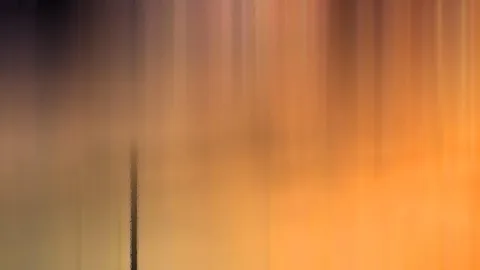 Vertikal verwischte Farbverläufe in Orange, Gelb und Dunkelbraun, oben dunkel, mit einer dünnen dunklen Linie links der Mitte.
