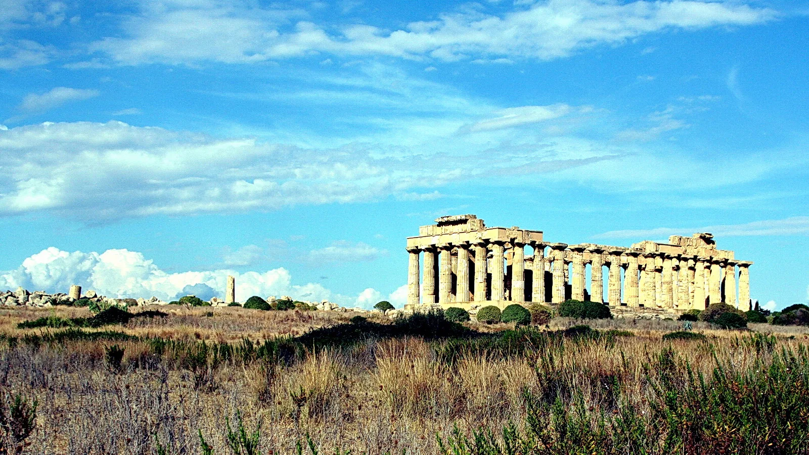 Antike griechische Tempelruine mit Säulenreihe über einer trockenen Graslandschaft unter blauem Himmel mit Wolken.
