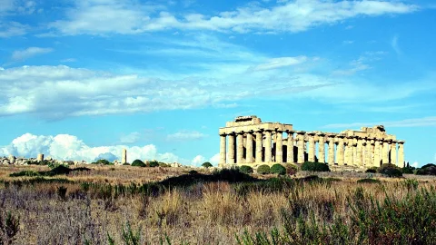 Antike griechische Tempelruine mit Säulenreihe über einer trockenen Graslandschaft unter blauem Himmel mit Wolken.