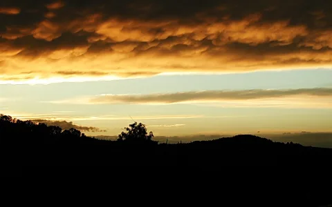 Dunkle Hügelsilhouette unter einem Himmel mit orangefarbenen und grauen Wolkenbändern bei Sonnenuntergang, links ein Baum.