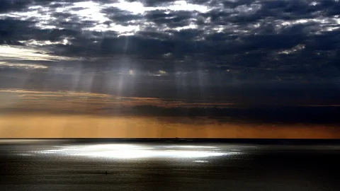 Dunkle Wolkendecke über einem ruhigen Meer, durch die Wolken fallen Sonnenstrahlen, auf dem Wasser glänzt ein helles Lichtfeld, am Horizont leuchtet ein orangefarbener Streifen.