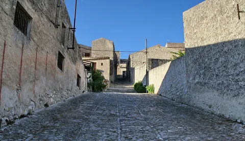 Schmale, steil ansteigende Gasse mit Kopfsteinpflaster zwischen hellen Steinmauern, am Ende führt eine Treppe nach oben, links ein vergittertes Fenster, blauer Himmel.