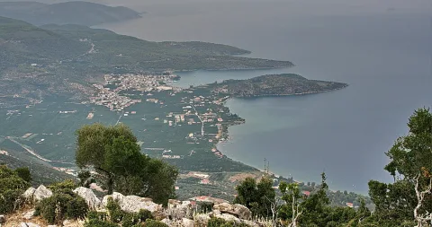 Blick von einem Bergrand auf ein Küstendorf mit Feldern, einer bewaldeten Halbinsel und dem blaugrauen Meer im Dunst.