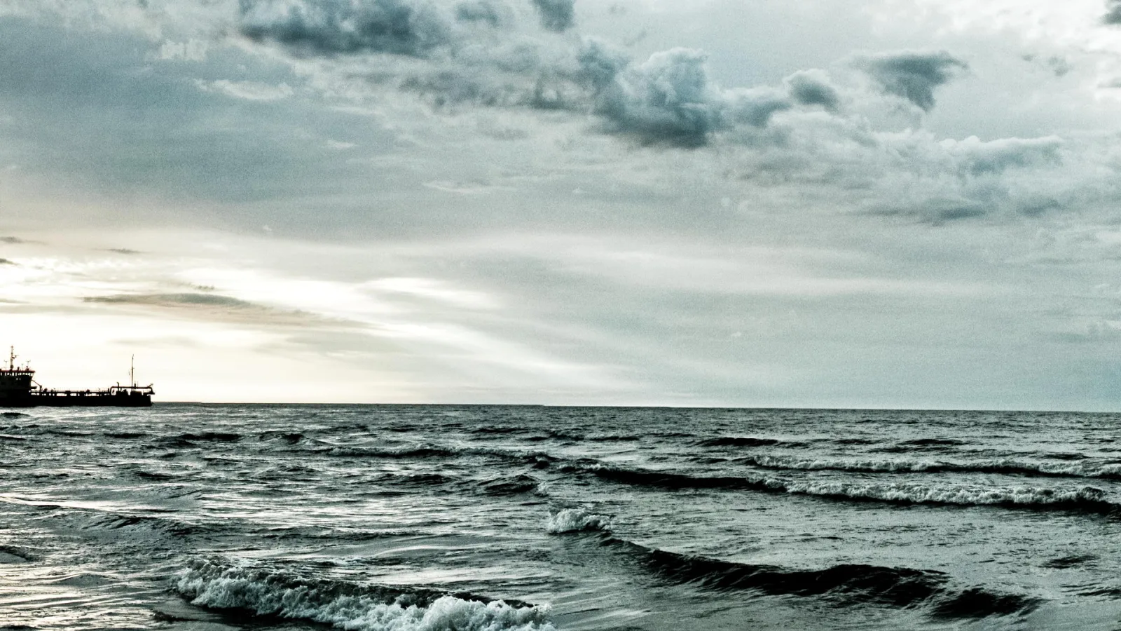 Graue Ostsee mit kurzen Wellen unter dichtem Wolkenhimmel, links am Horizont ein dunkler Frachter vor hellem Licht.