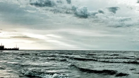 Graue Ostsee mit kurzen Wellen unter dichtem Wolkenhimmel, links am Horizont ein dunkler Frachter vor hellem Licht.