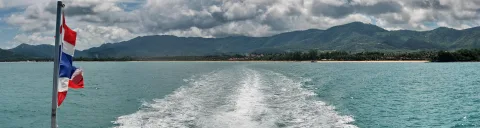 Kielwasser eines Bootes auf türkisfarbenem Meer, dahinter bewaldete Berge und ein heller Strand; am Heck weht eine rot-weiß-blaue Flagge.