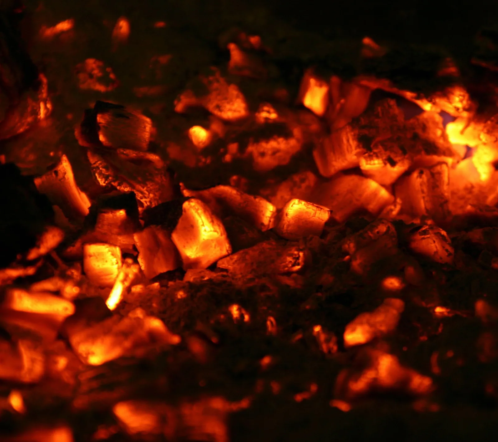 Nahaufnahme glühender Holzkohlestücke in Orange und Rot vor dunklem Hintergrund, teilweise mit heller Glut an den Kanten.